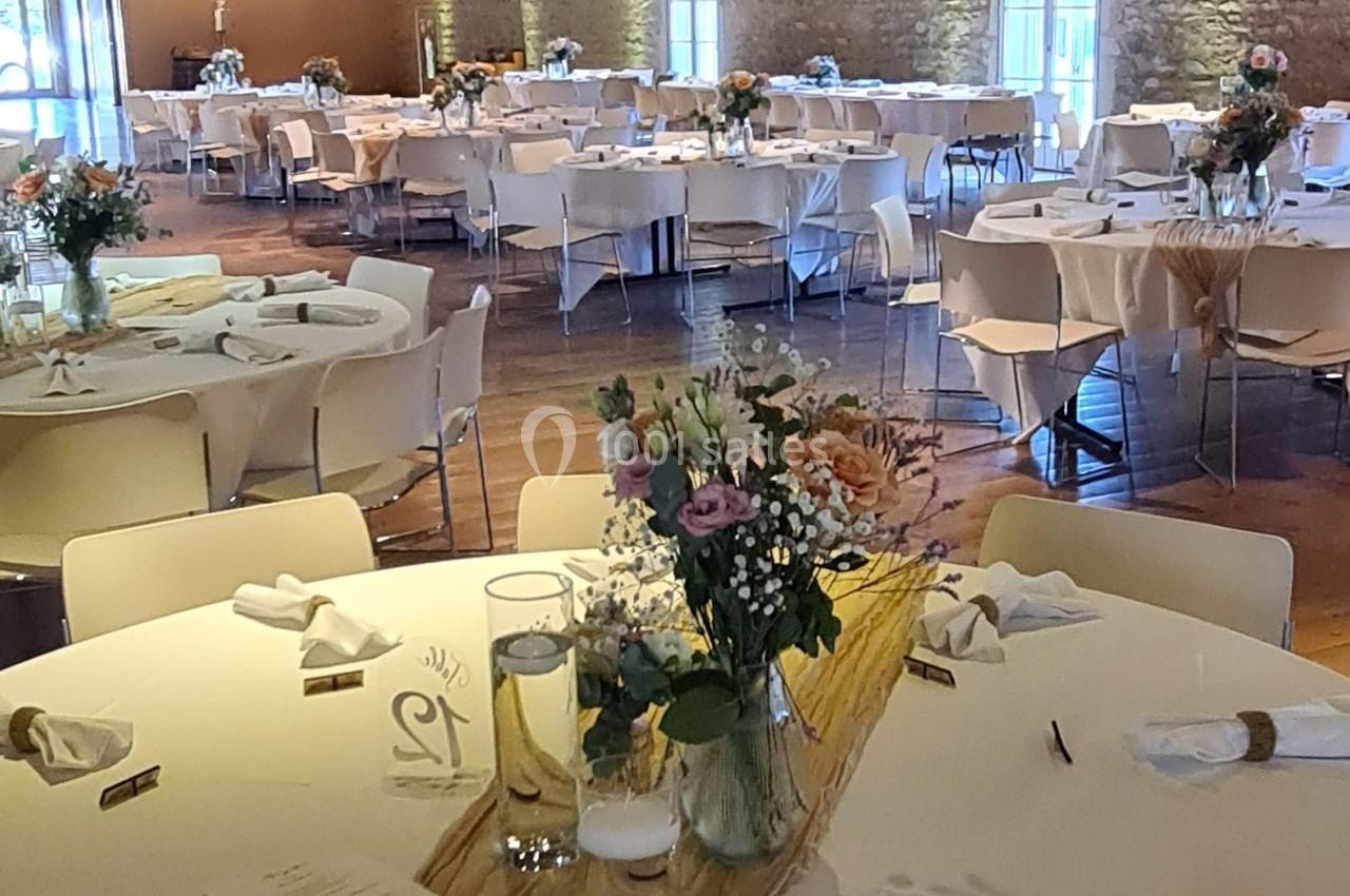 Salle de réception décorée avec des tables rondes, nappes blanches, centres de table fleuris et poutres apparentes au…