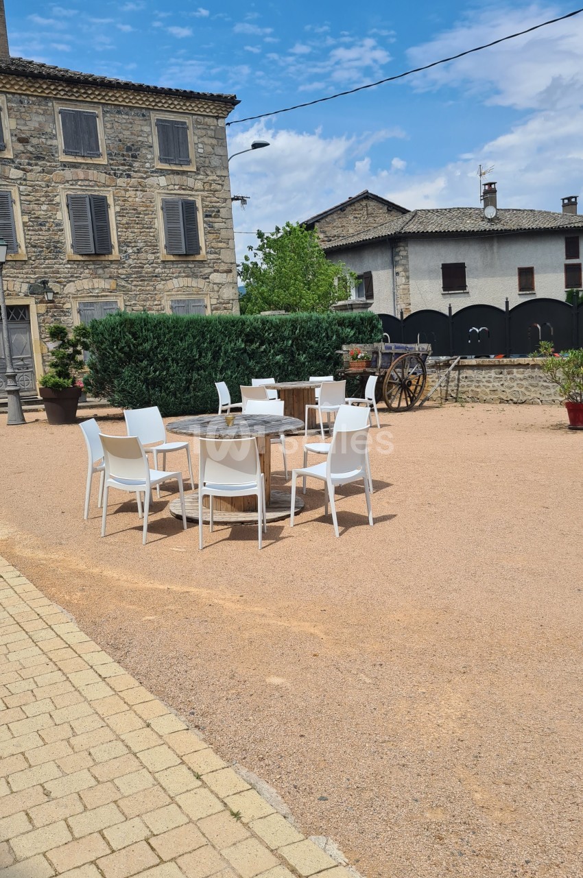Chaises blanches et tables en bois disposées sur une place en gravier devant des bâtiments en pierre.