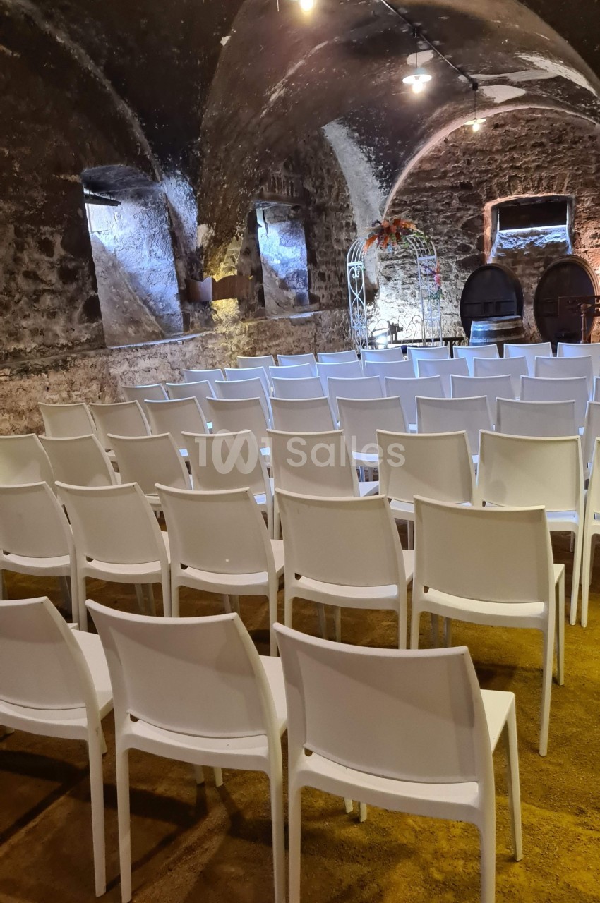 Salle voûtée en pierre avec des rangées de chaises blanches disposées face à une arche décorative.
