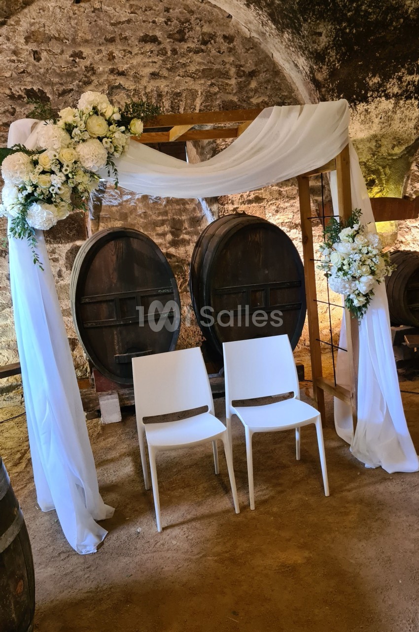 Arche décorée de fleurs blanches et de tissu, devant deux tonneaux en bois et deux chaises blanches, dans une cave voûtée.