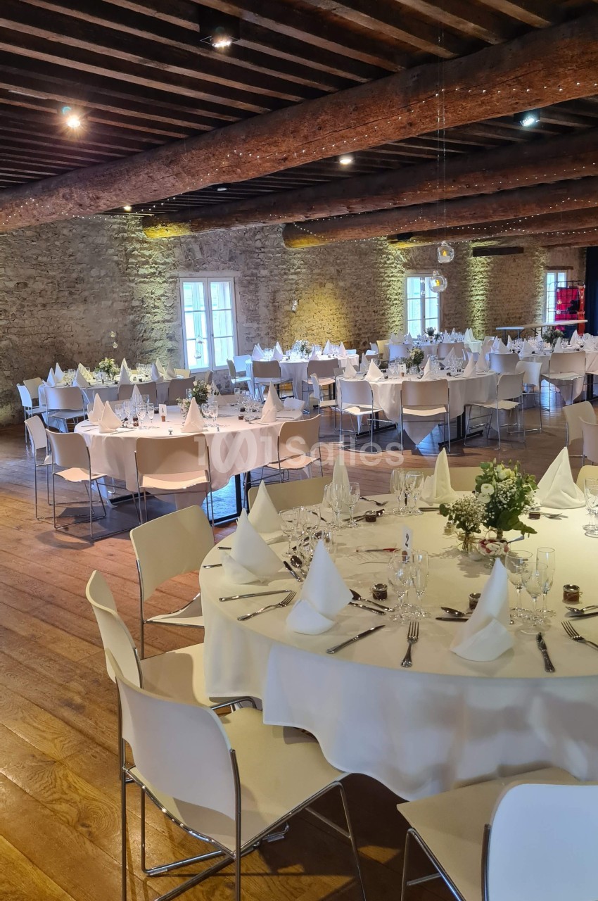Salle de réception avec tables rondes dressées, nappes blanches, chaises modernes et murs en pierre apparente.