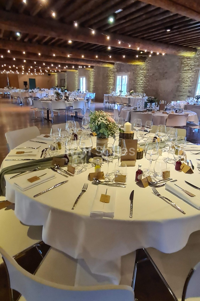 Salle de réception avec tables rondes dressées, décorées de fleurs, bougies et couverts élégants sous un plafond en bois.