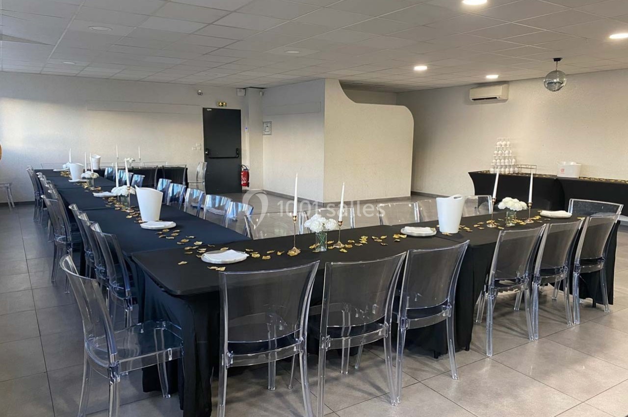 Salle aménagée avec deux longues tables noires décorées de bougies et pétales dorés, entourées de chaises transparentes.