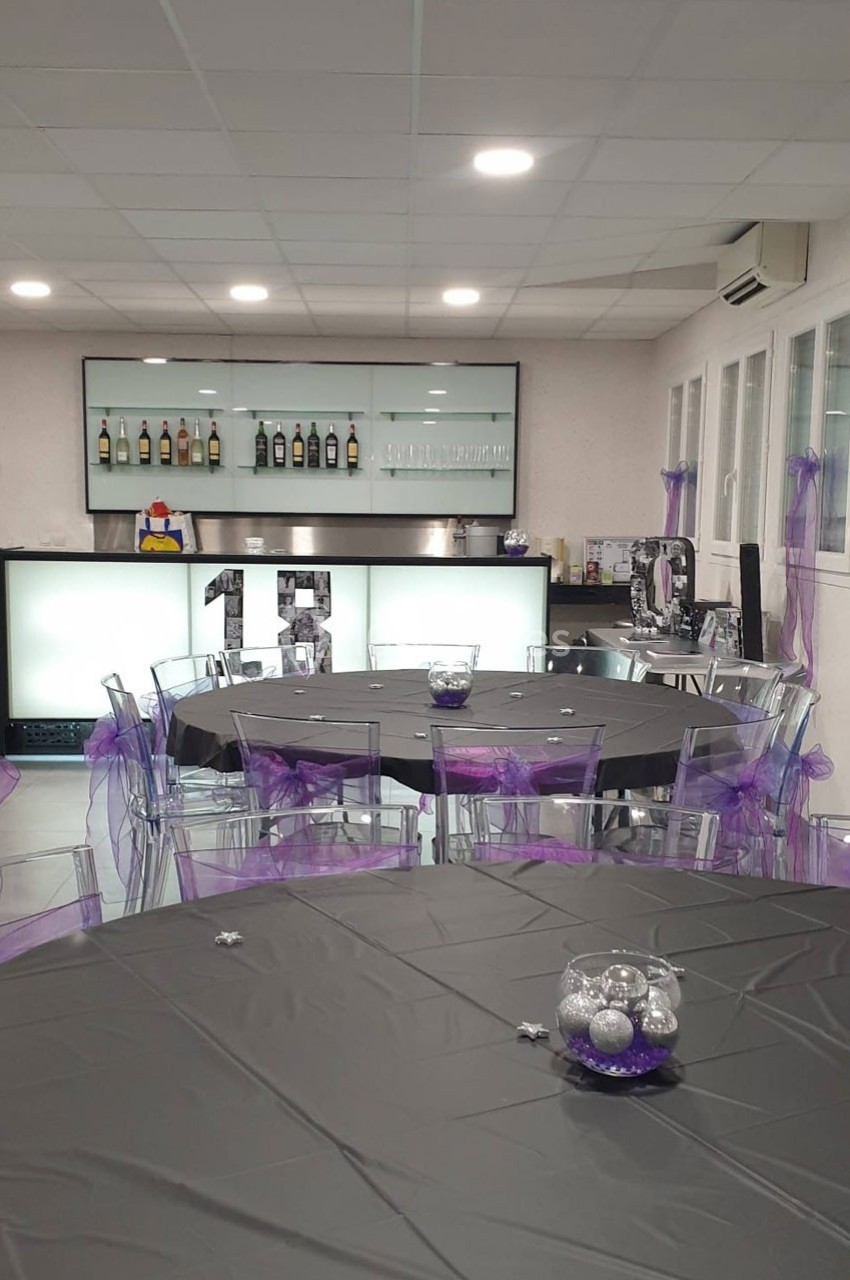 Salle décorée pour un événement avec tables rondes noires, chaises transparentes ornées de rubans violets et un bar en…