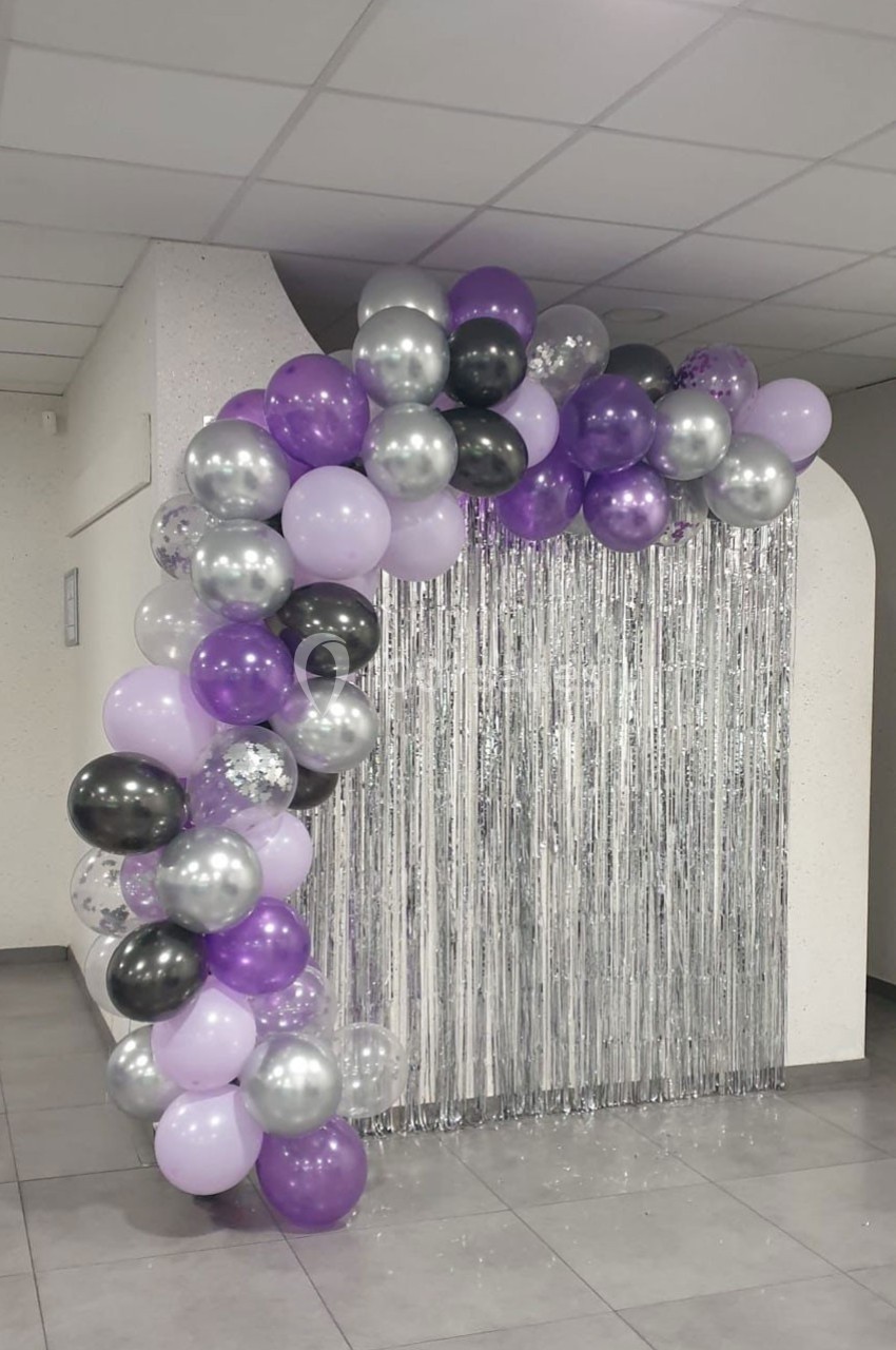 Guirlande de ballons violets, argentés et noirs encadrant un rideau argenté dans une salle au mur clair.