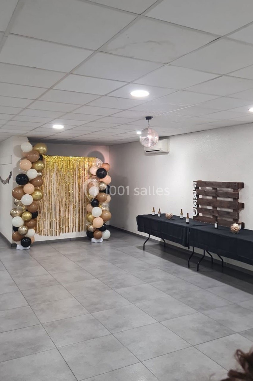 Décoration de fête avec arche de ballons beige, noir et or, rideau doré, table avec bouteilles et panneau en bois.