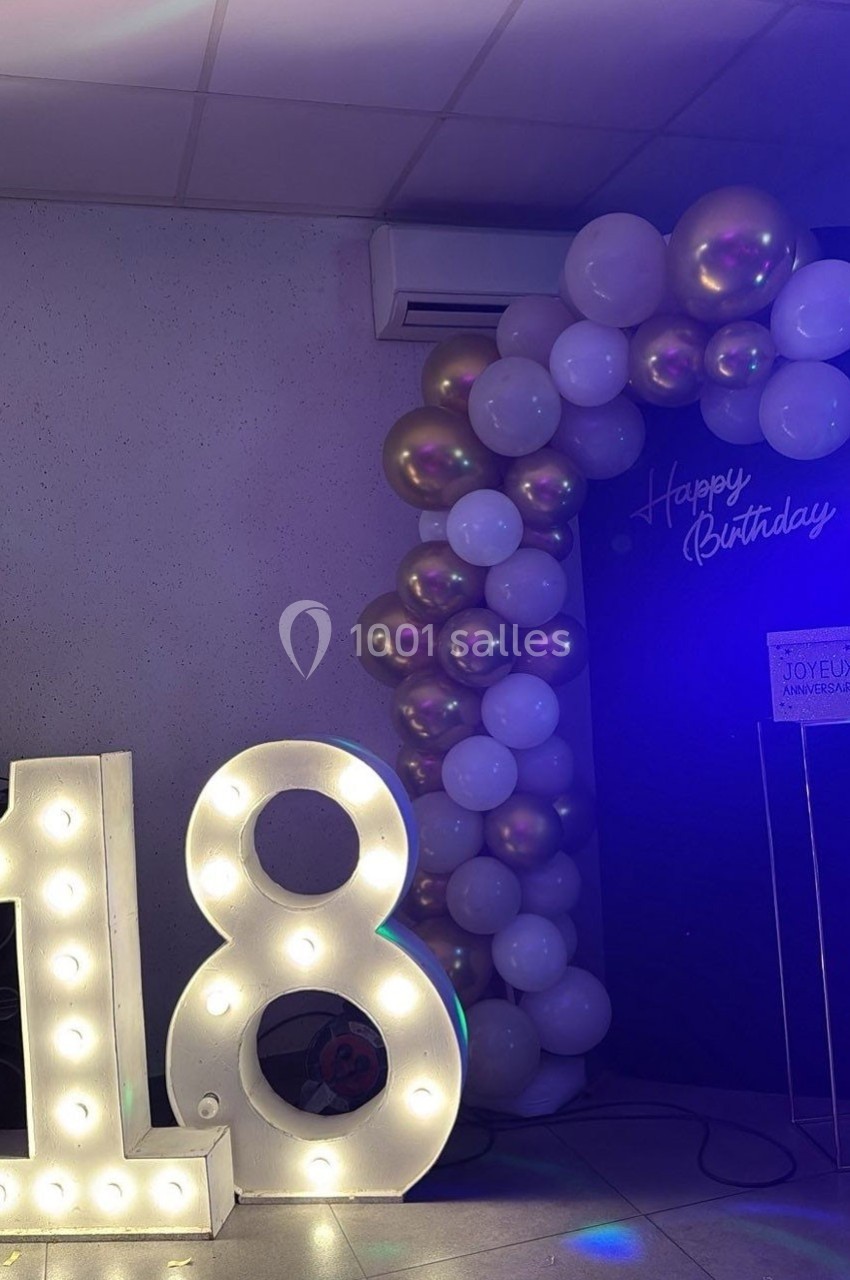 Décoration d'anniversaire avec un chiffre lumineux ’18’ et une arche de ballons blancs et dorés sous un panneau ’Happy…