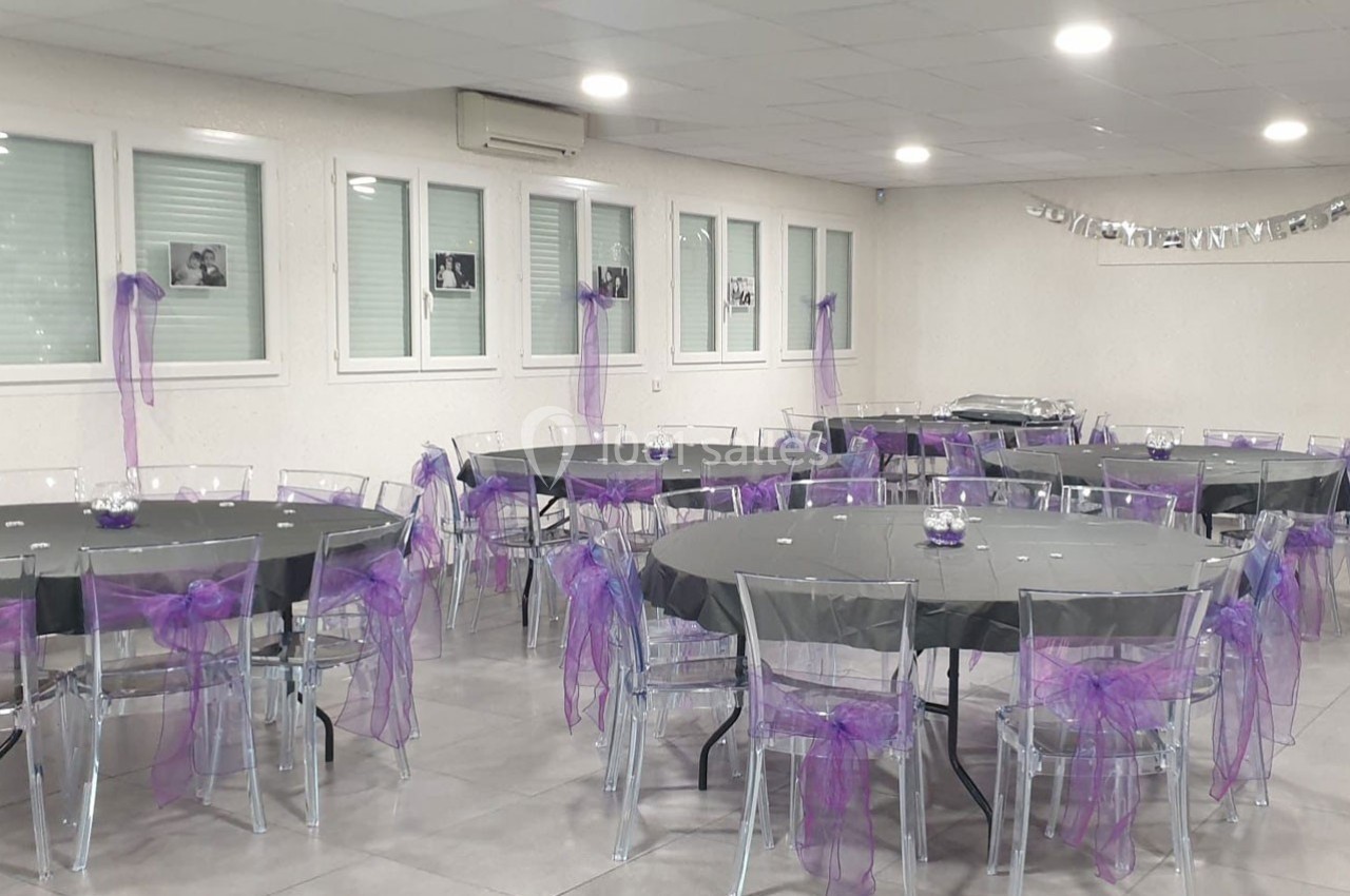 Salle décorée pour un événement avec des tables rondes nappées de noir et des chaises ornées de rubans violets.