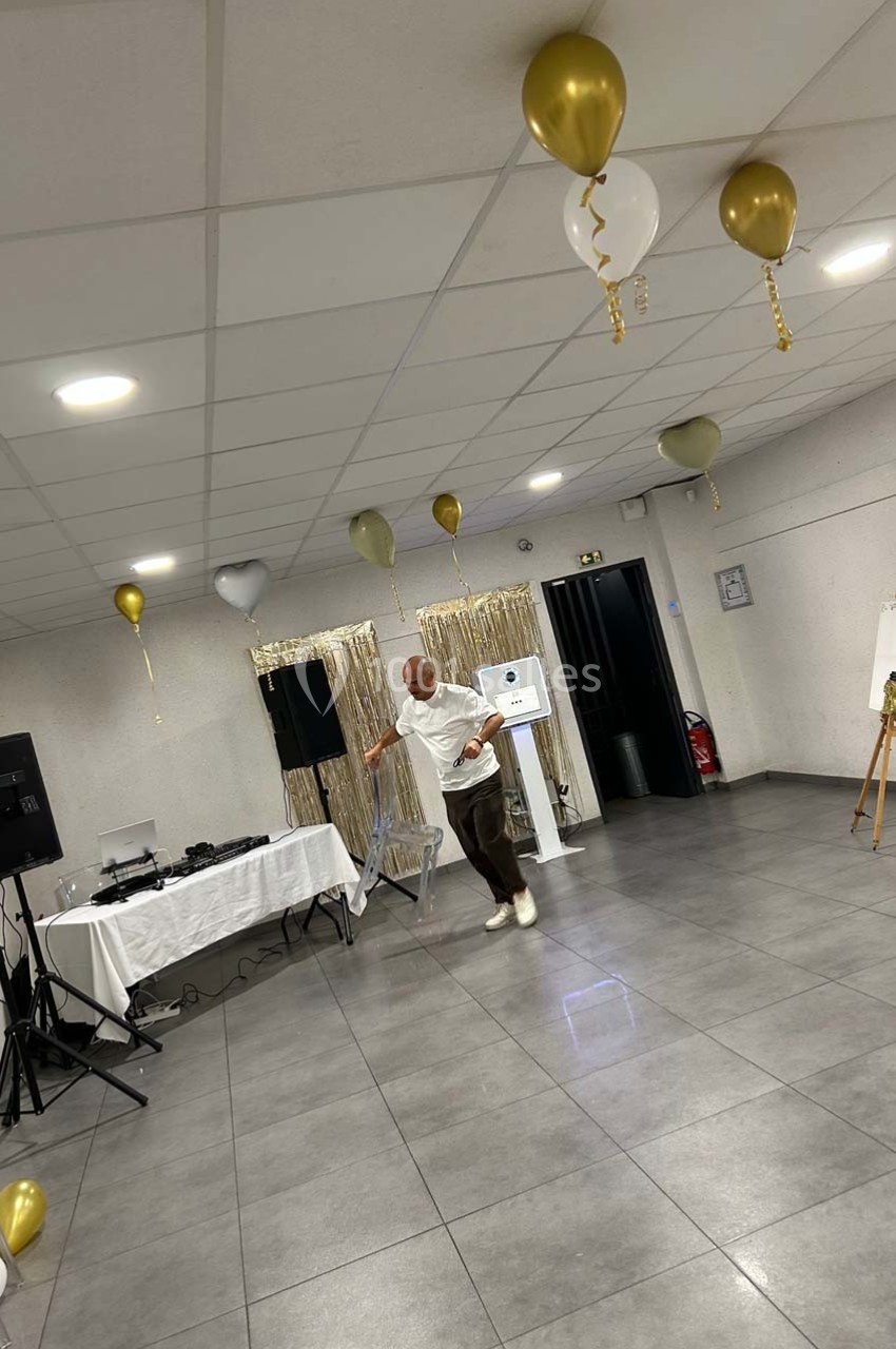 Un homme danse seul dans une salle décorée de ballons dorés et blancs, avec une table de DJ en arrière-plan.