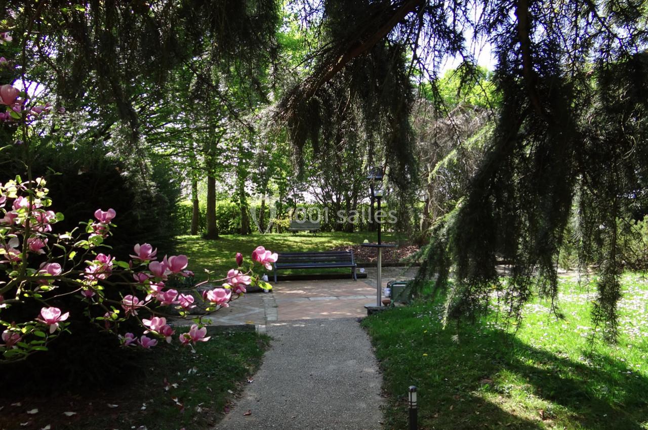 Location salle La Villeneuve-en-Chevrie (Yvelines) - La Grange des Mollières #3 Allée bordée de magnolias et d'arbres menant à un banc dans un parc verdoyant et ensoleillé.