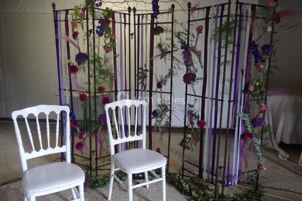 Location salle La Villeneuve-en-Chevrie (Yvelines) - La Grange des Mollières #9 Deux chaises blanches devant une structure métallique décorée de fleurs, feuillages et rubans violets.