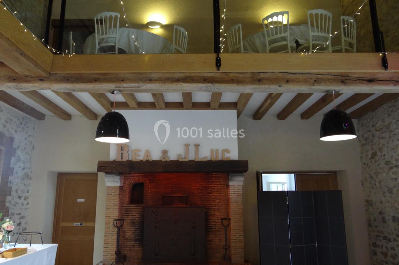 Location salle La Villeneuve-en-Chevrie (Yvelines) - La Grange des Mollières #10 Cheminée en briques dans une salle avec poutres apparentes, tables dressées à l'étage et lettres décoratives au mur.