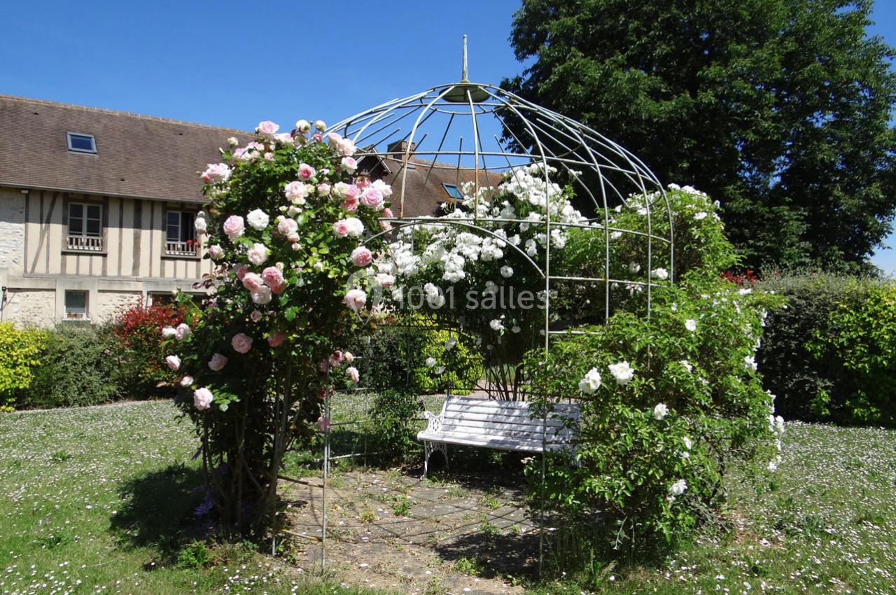 Location salle La Villeneuve-en-Chevrie (Yvelines) - La Grange des Mollières #24 Tonnelle en métal recouverte de rosiers fleuris, avec un banc blanc, dans un jardin ensoleillé près d'une maison à…