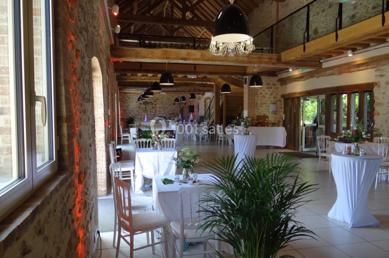 Location salle La Villeneuve-en-Chevrie (Yvelines) - La Grange des Mollières #29 Salle de réception lumineuse avec murs en pierre, poutres apparentes, tables décorées et grandes fenêtres.