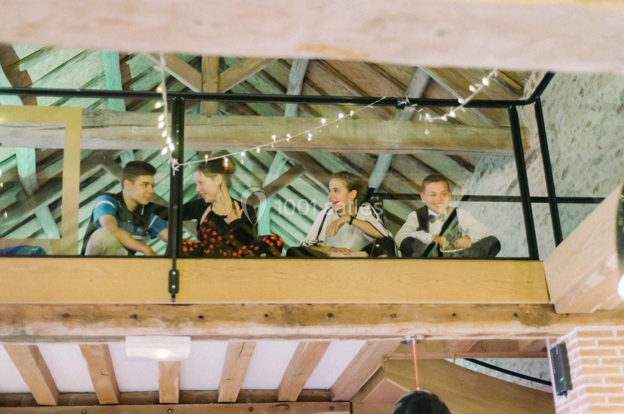 Location salle La Villeneuve-en-Chevrie (Yvelines) - La Grange des Mollières #11 Quatre personnes assises sur une mezzanine en bois, entourées de guirlandes lumineuses, dans un espace au plafond en poutres…