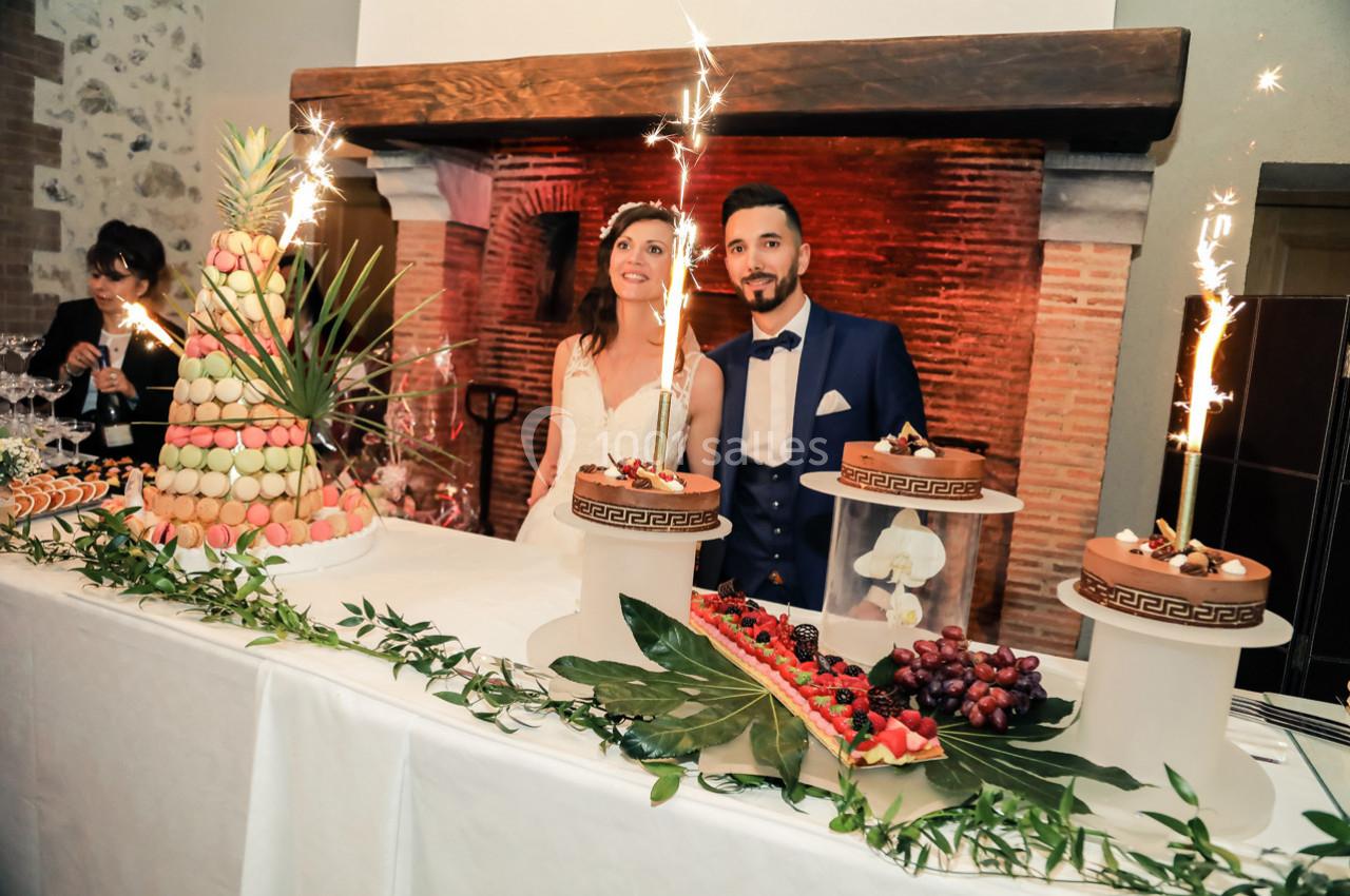 Location salle La Villeneuve-en-Chevrie (Yvelines) - La Grange des Mollières #4 Un couple souriant devant une table décorée avec des desserts, des macarons et des gâteaux ornés de cierges magiques.
