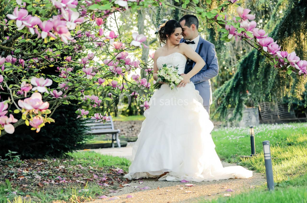 Location salle La Villeneuve-en-Chevrie (Yvelines) - La Grange des Mollières #25 Un couple de mariés pose dans un jardin fleuri, entouré de magnolias roses et d'une lumière douce.