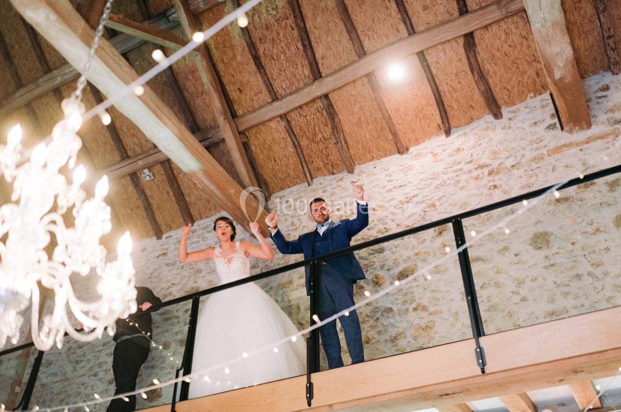 Location salle La Villeneuve-en-Chevrie (Yvelines) - La Grange des Mollières #28 Un couple en tenue de mariage se tient sur une mezzanine en bois, sous un plafond avec poutres apparentes et guirlandes…