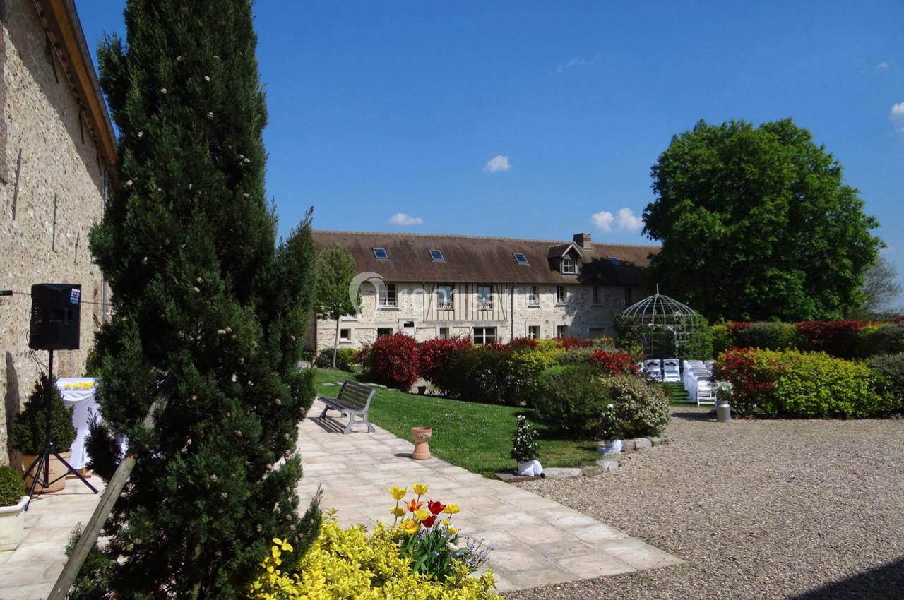 Location salle La Villeneuve-en-Chevrie (Yvelines) - La Grange des Mollières #2 Cour d'une propriété en pierre avec jardin fleuri, allée gravillonnée, et tonnelle sous un ciel bleu.