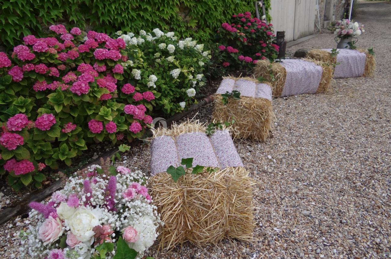 Location salle La Villeneuve-en-Chevrie (Yvelines) - La Grange des Mollières #32 Bottes de paille recouvertes de tissu rose, disposées en rangée, entourées de fleurs et de gravier.