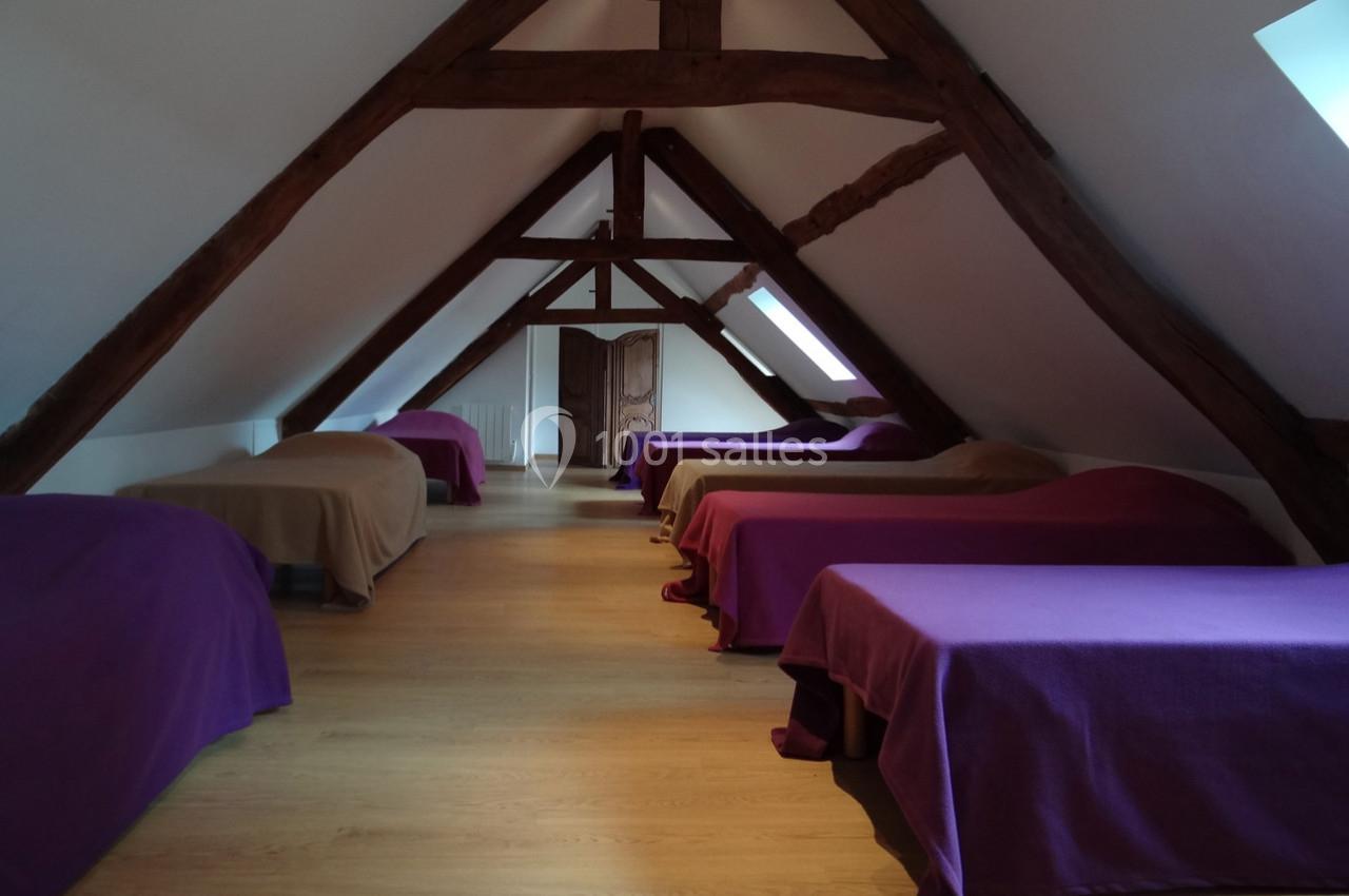 Location salle La Villeneuve-en-Chevrie (Yvelines) - La Grange des Mollières #51 Dortoir mansardé avec poutres apparentes, lits alignés recouverts de couvertures colorées, sol en bois clair.