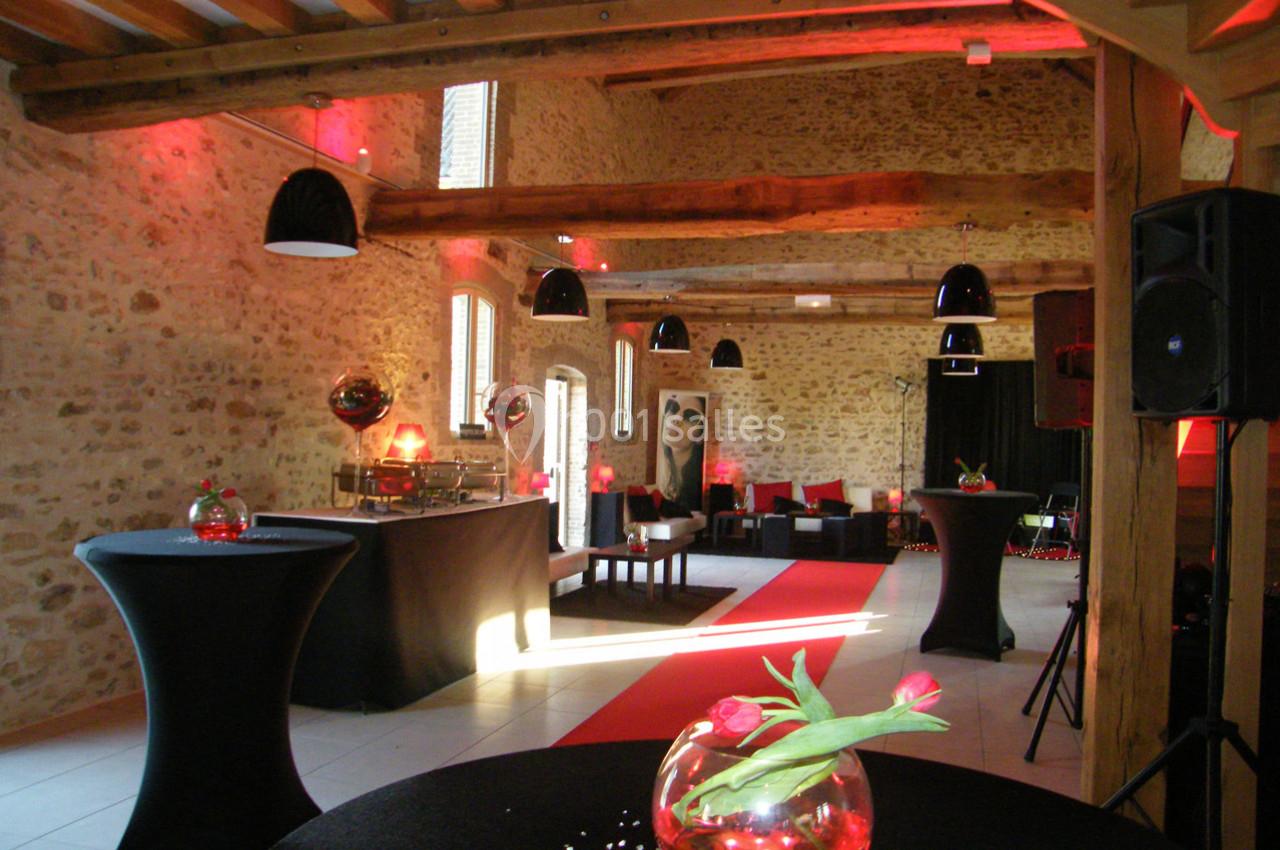 Location salle La Villeneuve-en-Chevrie (Yvelines) - La Grange des Mollières #17 Salle en pierre avec poutres apparentes, tables hautes décorées de fleurs, éclairage rouge et mobilier moderne.