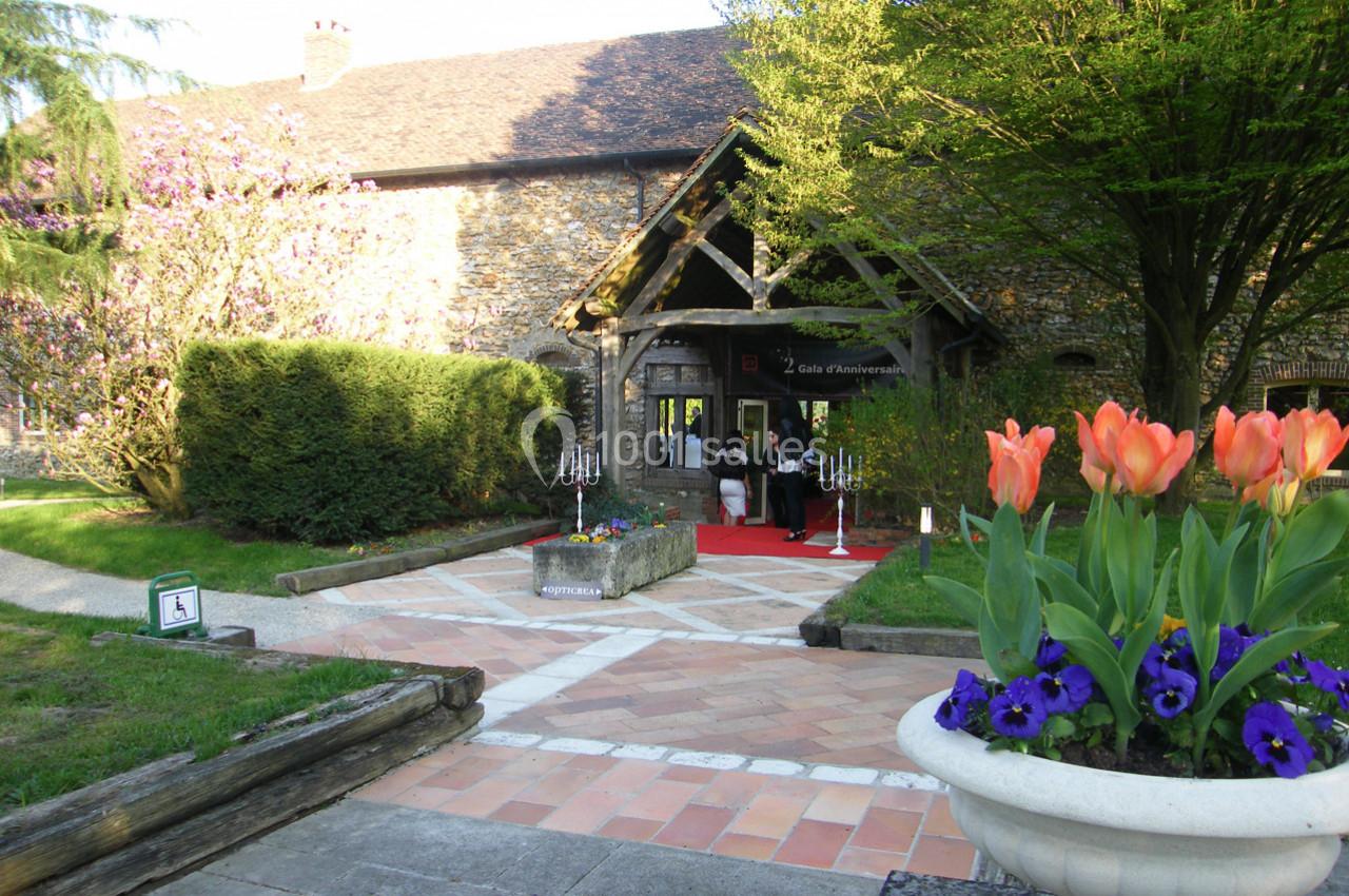 Location salle La Villeneuve-en-Chevrie (Yvelines) - La Grange des Mollières #16 Entrée d'un bâtiment en pierre avec un tapis rouge, entourée de verdure, de fleurs et d'arbres en arrière-plan.