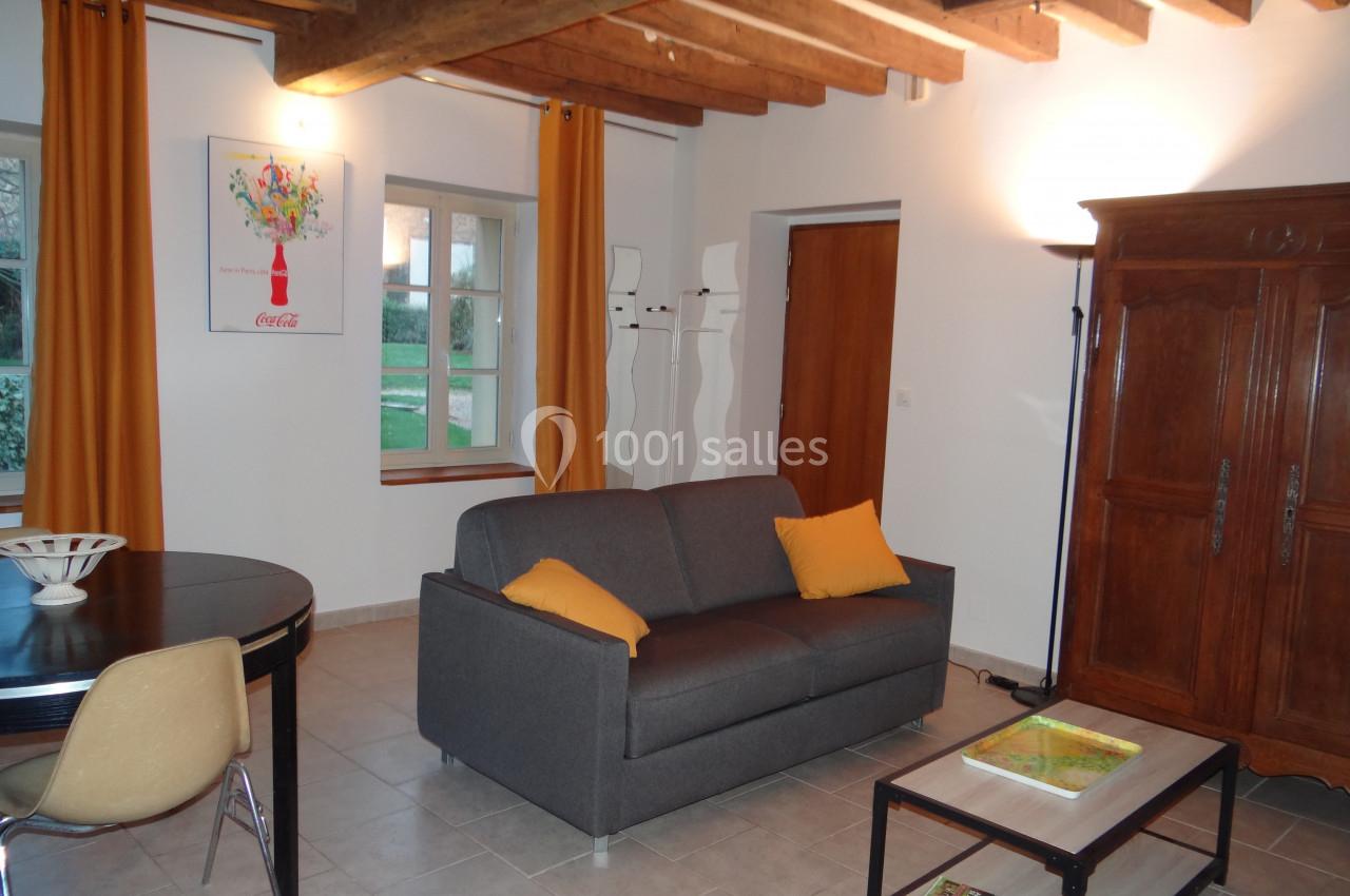 Location salle La Villeneuve-en-Chevrie (Yvelines) - La Grange des Mollières #54 Salon lumineux avec canapé gris, table basse, table à manger, poutres apparentes et rideaux jaunes.