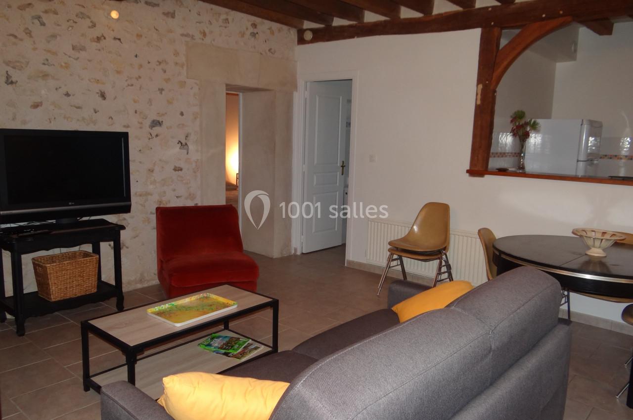 Location salle La Villeneuve-en-Chevrie (Yvelines) - La Grange des Mollières #55 Salon avec murs en pierre, canapé gris, fauteuil rouge, table ronde, télévision et cuisine ouverte en arrière-plan.