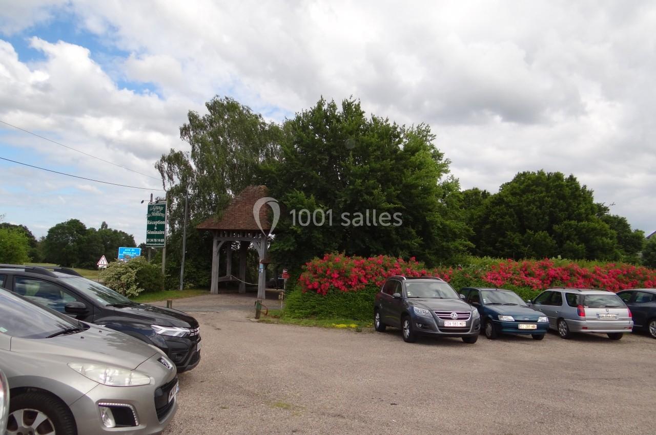Location salle La Villeneuve-en-Chevrie (Yvelines) - La Grange des Mollières #40 Parking extérieur avec plusieurs voitures, entouré de buissons fleuris et d'arbres sous un ciel partiellement nuageux.