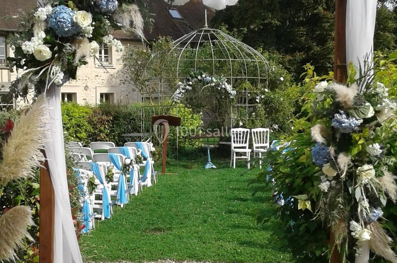 Location salle La Villeneuve-en-Chevrie (Yvelines) - La Grange des Mollières #46 Cérémonie en extérieur avec chaises décorées, arche florale et structure métallique dans un jardin verdoyant.