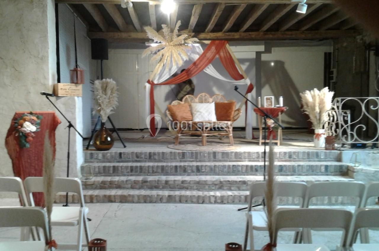 Location salle La Villeneuve-en-Chevrie (Yvelines) - La Grange des Mollières #47 Scène décorée avec des fauteuils en rotin, voilages rouges et blancs, et ornements de pampas, vue depuis des chaises…