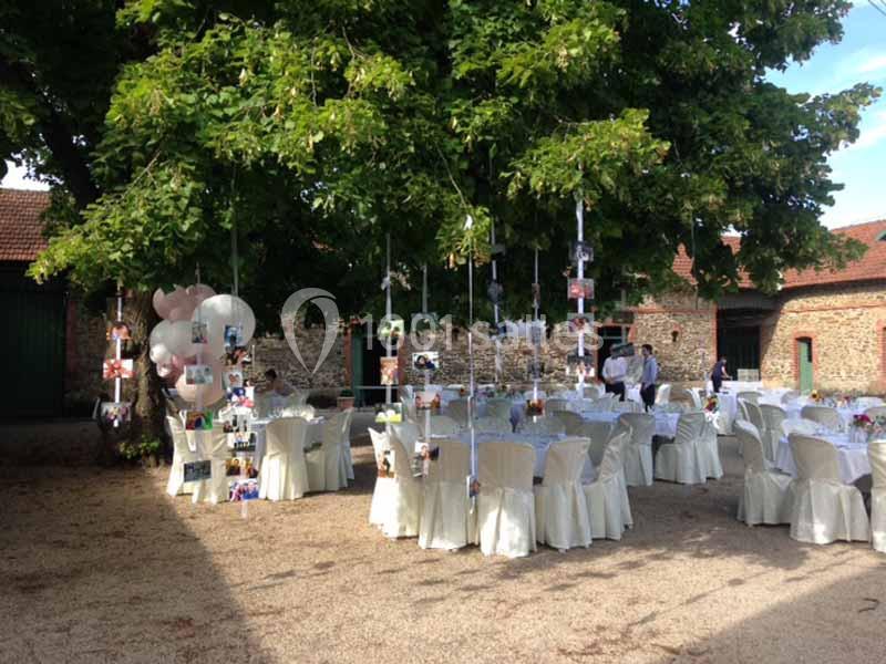 Location salle Haute-Goulaine (Loire-Atlantique) - La Cour des Montys #16 Tables décorées pour un événement en plein air sous un grand arbre, avec des photos suspendues et des chaises blanches.