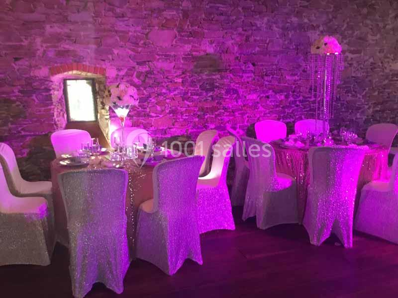 Location salle Haute-Goulaine (Loire-Atlantique) - La Cour des Montys #14 Salle décorée pour un événement avec des tables rondes, nappes scintillantes, chaises blanches et éclairage rose.