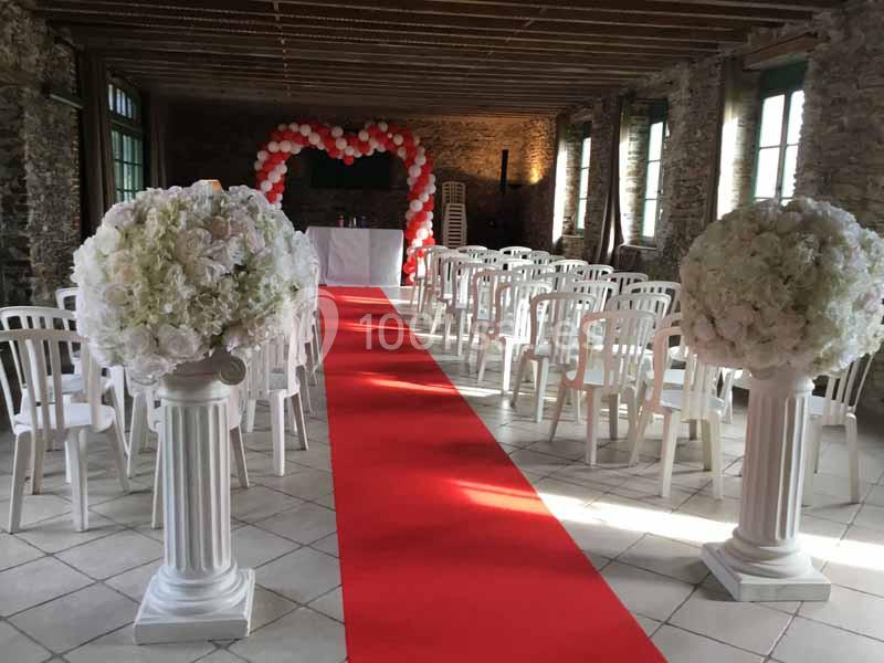 Location salle Haute-Goulaine (Loire-Atlantique) - La Cour des Montys #8 Salle décorée pour une cérémonie avec chaises blanches, arche de ballons rouges et blancs, et tapis rouge central.