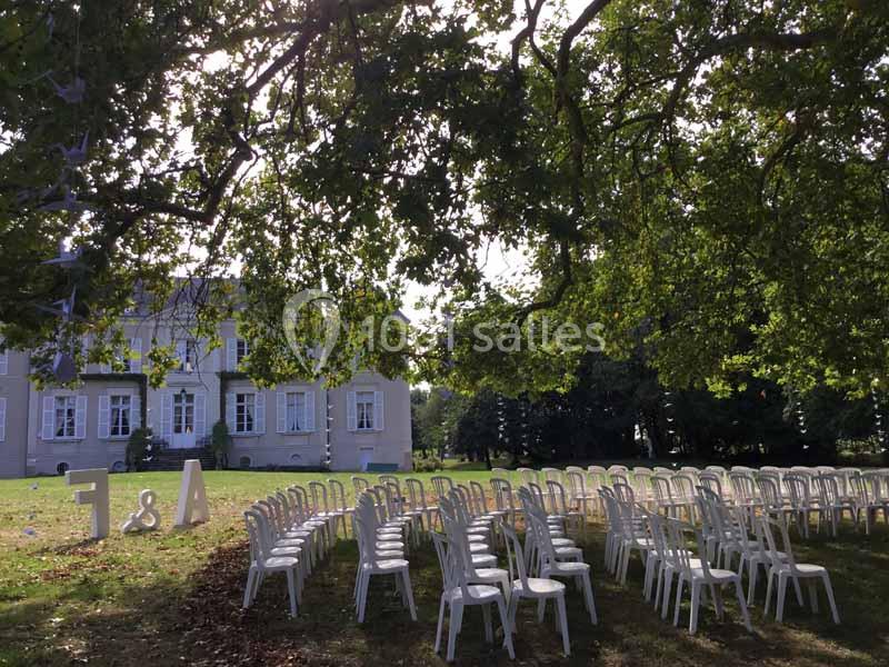 Location salle Haute-Goulaine (Loire-Atlantique) - La Cour des Montys #21 Chaises blanches disposées en rangées sur une pelouse ombragée devant un bâtiment ancien, préparées pour un événement.