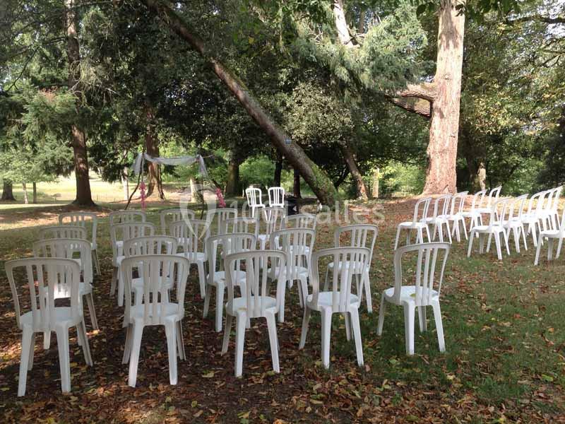 Location salle Haute-Goulaine (Loire-Atlantique) - La Cour des Montys #19 Chaises blanches en plastique disposées en rangées sur une pelouse ombragée, entourées d'arbres.