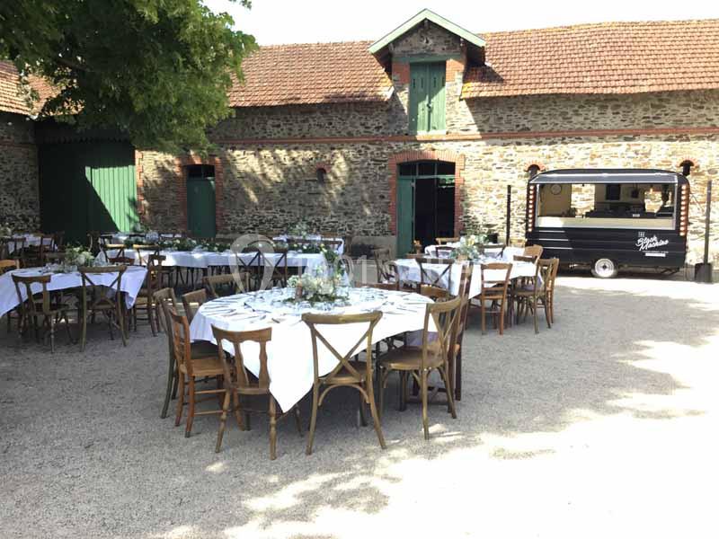 Location salle Haute-Goulaine (Loire-Atlantique) - La Cour des Montys #23 Tables rondes dressées avec nappes blanches dans une cour pavée, entourée de bâtiments en pierre et d'un food truck noir.