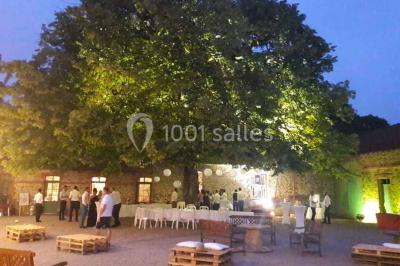 Miniature Location salle Haute-Goulaine (Loire-Atlantique) - La Cour des Montys #24 Cour extérieure éclairée en soirée, avec un grand arbre central, des tables, des palettes en bois et des invités debout.