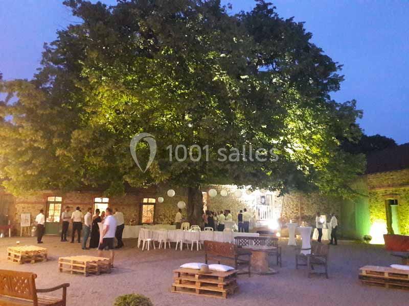 Location salle Haute-Goulaine (Loire-Atlantique) - La Cour des Montys #24 Cour extérieure éclairée en soirée, avec un grand arbre central, des tables, des palettes en bois et des invités debout.
