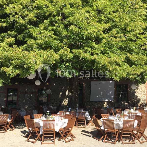 Location salle Haute-Goulaine (Loire-Atlantique) - La Cour des Montys #22 Tables dressées avec nappes blanches et chaises en bois disposées sous un grand arbre dans une cour ensoleillée.
