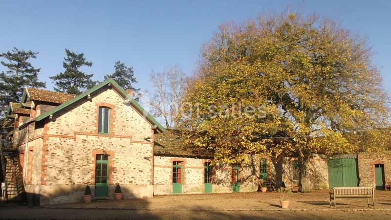 Location salle Haute-Goulaine (Loire-Atlantique) - La Cour des Montys #15 Bâtiment en briques rouges avec des volets verts, entouré d'arbres et baigné par une lumière ensoleillée.