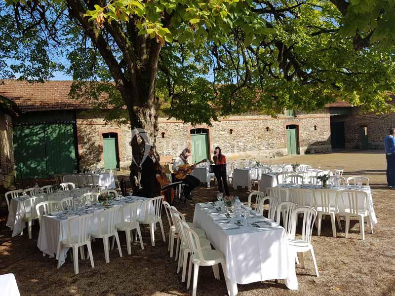 Location salle Haute-Goulaine (Loire-Atlantique) - La Cour des Montys #17 Tables dressées sous un grand arbre dans une cour ensoleillée, avec des musiciens jouant près des bâtiments en pierre.
