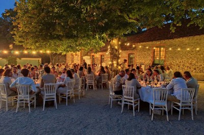 Un groupe de personnes rassemblées sous de grands arbres devant des bâtiments en pierre par une journée ensoleillée.