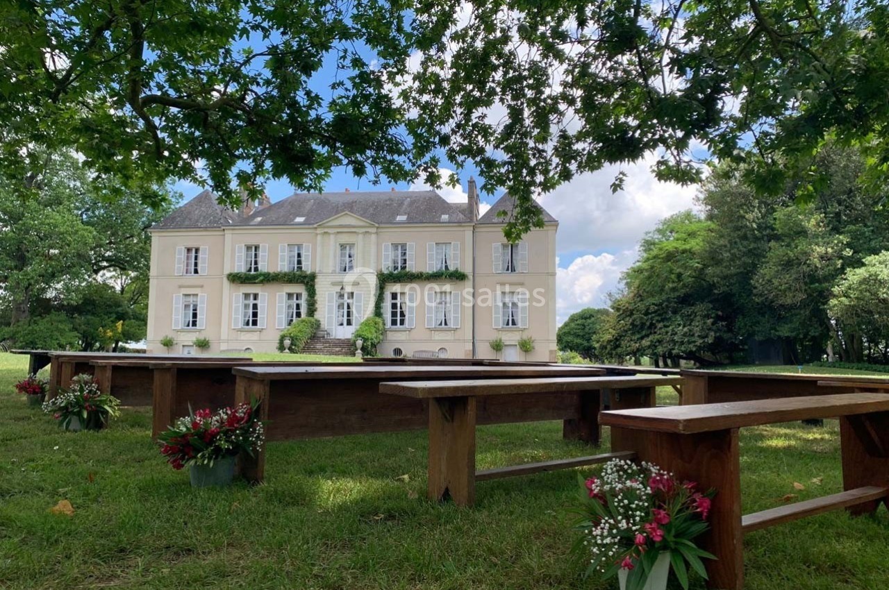 Façade d'un château clair entouré de verdure, avec des bancs en bois et des bouquets de fleurs au premier plan.