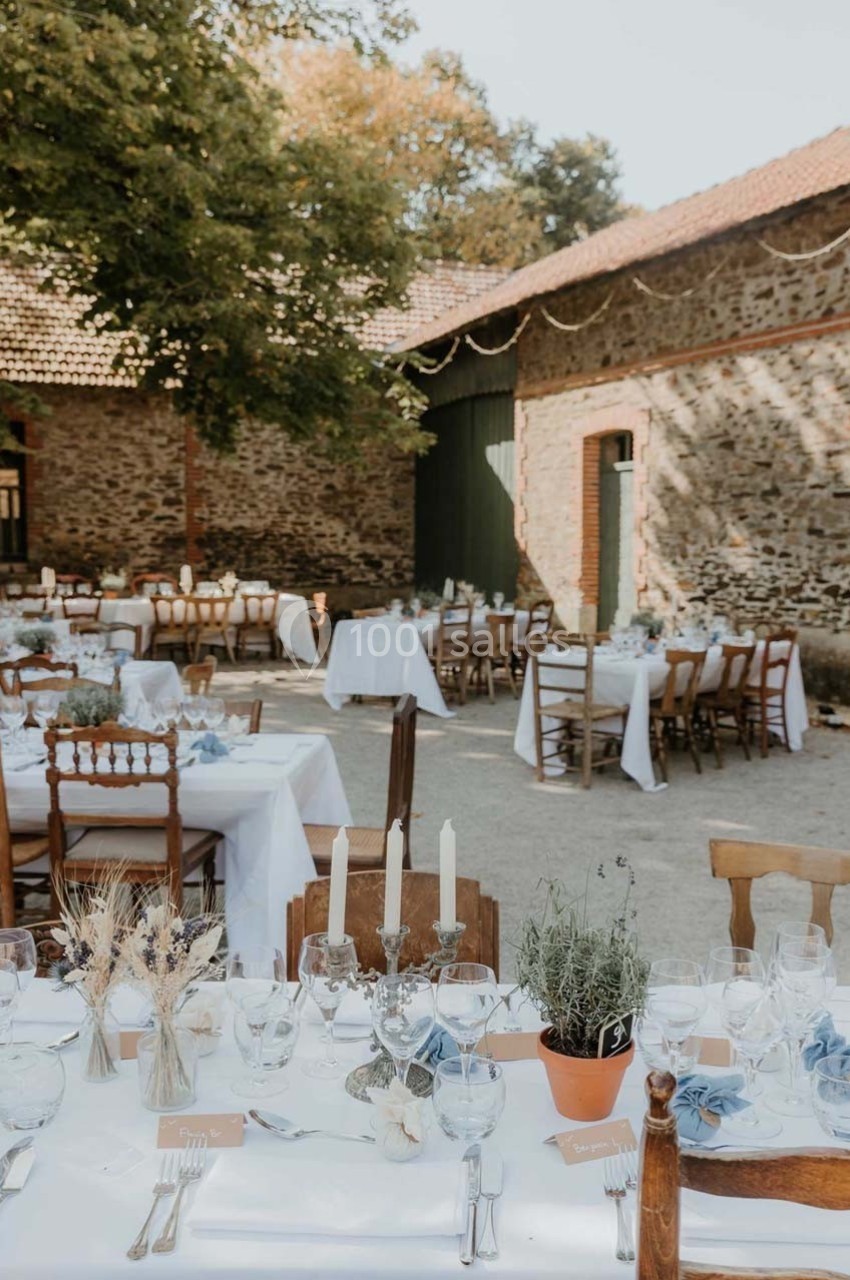 Tables dressées pour un repas en extérieur dans une cour pavée, entourée de bâtiments en pierre et d'arbres.