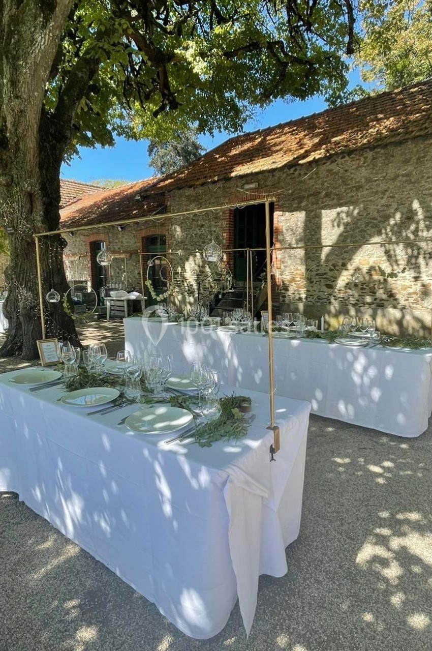 Tables dressées avec nappes blanches, vaisselle et décorations, installées en extérieur sous des arbres près d'un bâtiment…