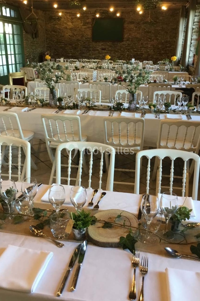 Salle de réception décorée avec des tables alignées, nappes blanches, chaises en bois clair et ornements végétaux.