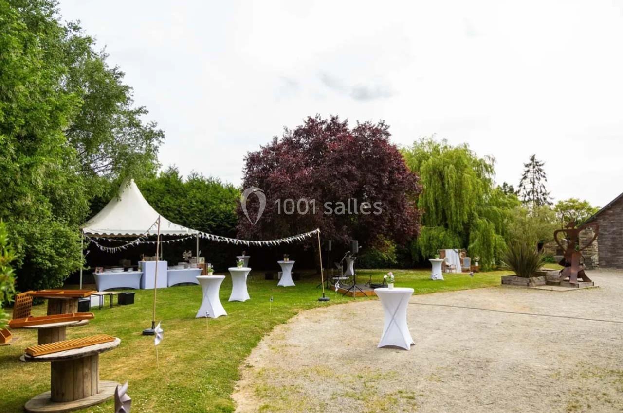 Jardin aménagé pour un événement avec tables hautes, tente blanche et décorations sous des arbres.