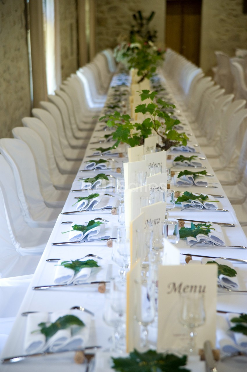 Table de banquet élégamment dressée avec nappes blanches, menus, couverts alignés et décoration de feuilles vertes.
